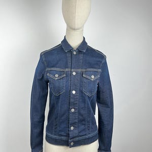 Peut inclure: Veste en jean bleu foncé avec une fermeture boutonnée, deux poches de poitrine et des manches longues. La veste a des boutons argentés et des coutures de couleur claire. La veste est présentée sur un mannequin.