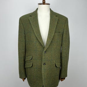 Puede incluir: Blazer de tweed verde con cierre de dos botones, solapas con muescas y dos bolsillos delanteros con solapa. La chaqueta se exhibe en un maniquí. La tela tiene un sutil estampado a cuadros.