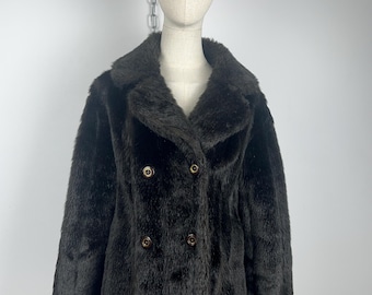 May include: A dark brown faux fur coat with a double-breasted design. The coat features a wide collar and four gold-colored buttons. The coat is displayed on a mannequin.