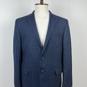 Puede incluir: Un blazer azul marino exhibido en un maniquí. El blazer tiene cierre de dos botones, solapa con muescas y dos bolsillos delanteros. La tela parece ser una mezcla de lana texturizada. El blazer es de estilo clásico, adecuado para diversas ocasiones.