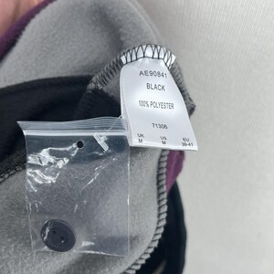 May include: Close-up of a black and gray fleece garment with a purple accent. A tag reads "AE90841 BLACK 100% POLYESTER". A small plastic bag contains a black button. The garment is medium size.