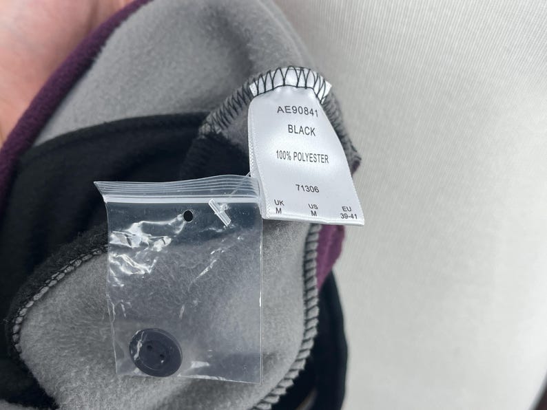 May include: Close-up of a black and gray fleece garment with a purple accent. A tag reads "AE90841 BLACK 100% POLYESTER". A small plastic bag contains a black button. The garment is medium size.