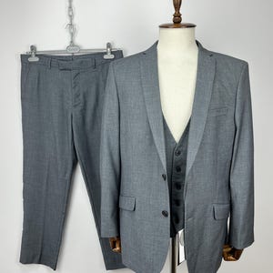 May include: A three-piece gray suit displayed on a mannequin. The suit includes a blazer, vest, and trousers. The blazer has two front pockets and a two-button closure. The trousers are hanging on a hanger. The suit is made of a textured fabric.