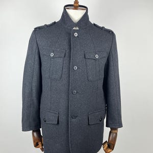 May include: A dark grey wool coat with a high collar and button-down front. The coat has two chest pockets and two lower pockets, all with button closures. Epaulettes are on the shoulders. The coat is displayed on a mannequin.