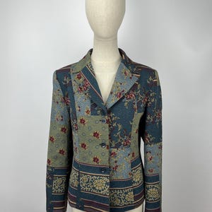 May include: A vintage blazer with a patchwork design featuring floral and geometric patterns in shades of teal, olive green, and burgundy. The blazer has a notched collar, three buttons, and long sleeves. The garment is displayed on a mannequin.