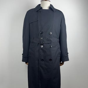 Puede incluir: Un abrigo tipo trench negro con diseño de doble botonadura, cinturón en la cintura y detalles de botones. El abrigo se exhibe en un maniquí, mostrando su estilo clásico y ajuste a medida. Una prenda de abrigo atemporal.