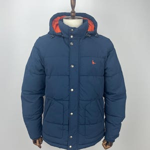 May include: A navy blue puffer jacket with an orange interior lining and a hood. The jacket has a button-up front, two front pockets, and an orange embroidered logo on the chest. The jacket is displayed on a mannequin.