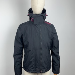 Könnte beinhalten: Schwarze Kapuzenjacke mit durchgehendem Reißverschluss. Die Jacke hat einen hohen Kragen und eine Kapuze mit rosa Akzenten. Der Markenname ist in Rosa auf der Schulter aufgedruckt. Die Jacke wird auf einer Schaufensterpuppe präsentiert.