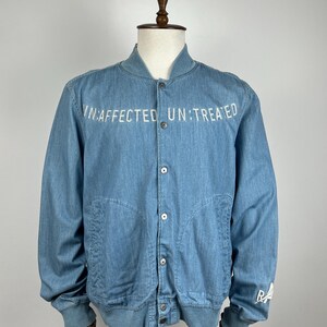 May include: Light blue denim bomber jacket with the text "UN:AFFECTED UN:TREATED" across the chest. The jacket has a button-up front, two front pockets, and ribbed cuffs and hem. The sleeve has the letters "RA" in white.