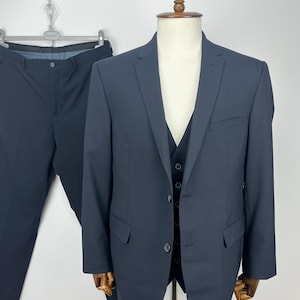 May include: A navy blue three-piece suit displayed on a mannequin. The suit includes a blazer, vest, and trousers. The blazer has two front pockets and a two-button closure. The trousers are hanging on a metal hanger. The suit is made of a solid, dark blue fabric.