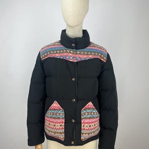 Puede incluir: Chaqueta acolchada negra con canesú y detalles de bolsillo estampados. La chaqueta presenta un cuello alzado, cierre de botones y dos bolsillos delanteros con un diseño geométrico en rosa, azul y beige.