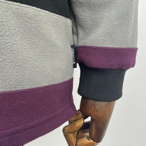 May include: Close-up of a fleece jacket with color-blocked panels in gray, black, and purple. The cuff is black with a purple trim. The Guinness logo is visible on the jacket.