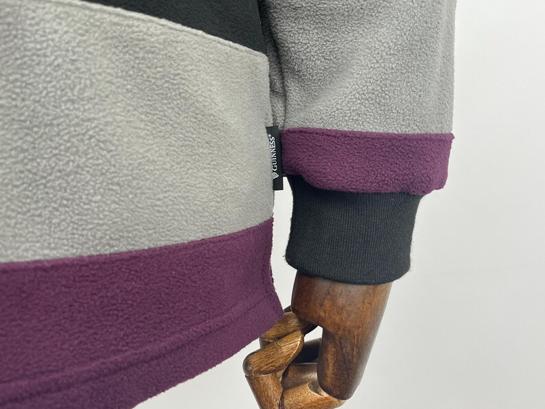 May include: Close-up of a fleece jacket with color-blocked panels in gray, black, and purple. The cuff is black with a purple trim. The Guinness logo is visible on the jacket.