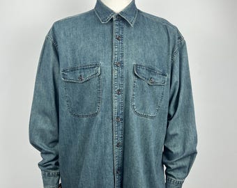 May include: A long-sleeved denim shirt in a medium blue wash, displayed on a mannequin. The shirt features two front pockets with button closures, a classic collar, and buttoned cuffs. The fabric has a slightly faded appearance.