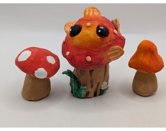 Conjunto de três figuras de cogumelos de argila feitas à mão, decoração campestre lúdica em tons de vermelho e laranja.