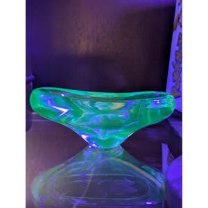 Puede incluir: Un cuenco de vidrio verde brillante con una base única en forma de reloj de arena. El cuenco tiene un diseño suave y curvado y refleja la luz, creando una vibrante exhibición de color. El vidrio parece ser una pieza vintage.