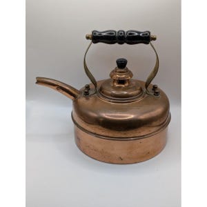 May include: A copper tea kettle with a black wooden handle and a spout. The kettle has a round body and a lid with a black knob. The handle is attached to the kettle with brass-colored metal supports. The kettle is likely vintage.