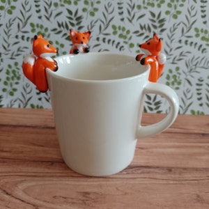 Peut inclure: Une tasse en céramique blanche ornée de trois figurines de renards orange et blanc. Les renards ont des yeux noirs et sont placés sur le bord. La tasse est posée sur une surface en bois, avec un motif de feuilles vertes et blanches en arrière-plan.