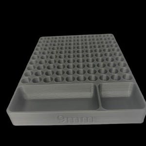 May include: A gray 9mm ammunition tray with multiple circular holes and two rectangular compartments. The tray is designed to hold and organize ammunition. The text "9mm" is visible on the front of the tray.