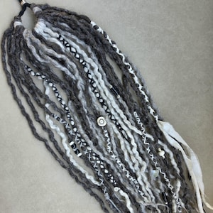 May include: A set of synthetic hair extensions featuring a mix of dark gray, light gray, and white yarn-like strands. The extensions are attached to a black elastic band and decorated with silver-toned metal charms, beads, and white fabric ribbons.
