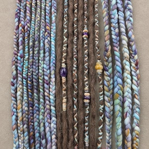 May include: A set of hair extensions featuring a mix of braided strands in variegated shades of blue, purple, and cream, alongside brown dreadlocks wrapped with colorful thread and accented with small, smooth, polished stone beads.