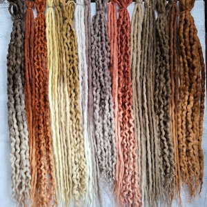 May include: A collection of long, textured synthetic dreadlocks arranged in a vertical row. The strands feature a diverse colour palette including shades of dark brown, copper, golden yellow, cream, white, soft grey, dusty rose, and tan.