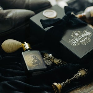 A bottle of The Yellow Wallpaper perfume with its haunting illustrated label lies on black velvet beside its ornate gift box, surrounded by antique trinkets, a pearl-topped clasp, and worn Victorian boots. The dim light and sepia tones cast the scene like a forgotten relic from a gothic boudoir, steeped in unease and decadent decay.