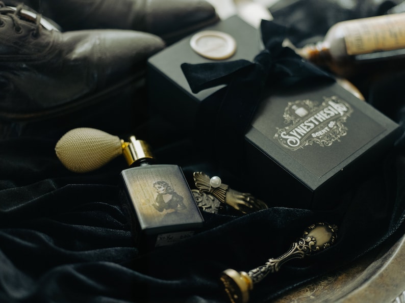 A bottle of The Yellow Wallpaper perfume with its haunting illustrated label lies on black velvet beside its ornate gift box, surrounded by antique trinkets, a pearl-topped clasp, and worn Victorian boots. The dim light and sepia tones cast the scene like a forgotten relic from a gothic boudoir, steeped in unease and decadent decay.