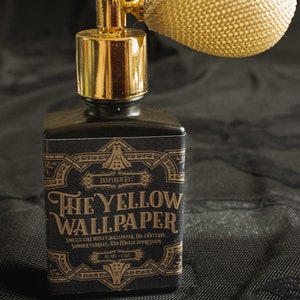 A square black perfume bottle labeled “The Yellow Wallpaper” features a gold atomizer with an ivory netted bulb, standing starkly against black patterned fabric. The dramatic light and shadow heighten the tension, casting the scent as something unsettlingly elegant—an object that whispers of confinement, decay, and beauty pressed beneath a heavy atmosphere.