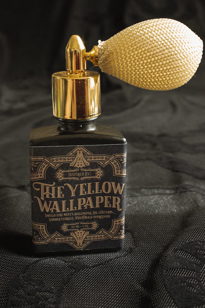 A square black perfume bottle labeled “The Yellow Wallpaper” features a gold atomizer with an ivory netted bulb, standing starkly against black patterned fabric. The dramatic light and shadow heighten the tension, casting the scent as something unsettlingly elegant—an object that whispers of confinement, decay, and beauty pressed beneath a heavy atmosphere.