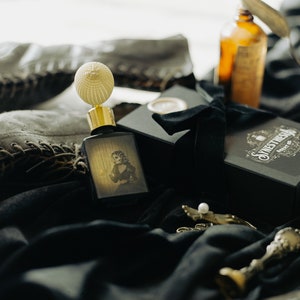 The Yellow Wallpaper perfume bottle, with its spectral illustrated label, rests on draped black velvet beside its ornate gift box, vintage lace-up boots, and an amber apothecary bottle. The pale light softens the scene, giving the objects the fragile, ghostly quality of relics left behind in a long-abandoned room.