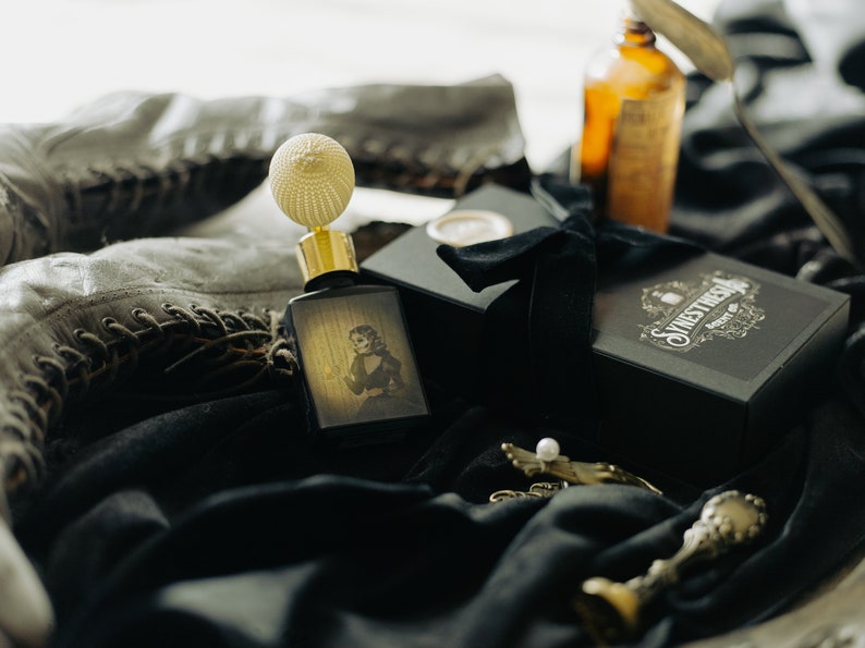 The Yellow Wallpaper perfume bottle, with its spectral illustrated label, rests on draped black velvet beside its ornate gift box, vintage lace-up boots, and an amber apothecary bottle. The pale light softens the scene, giving the objects the fragile, ghostly quality of relics left behind in a long-abandoned room.