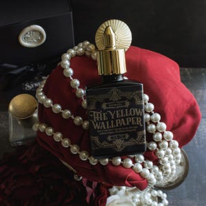 A bottle of “The Yellow Wallpaper” perfume with a gold atomizer sits atop deep red fabric, draped in pearls and surrounded by dark red and white roses. The composition, dramatic with shadows and heavy textures, evokes a sense of trapped opulence—luxury slipping into confinement, as if the beauty itself carries a fevered unease.