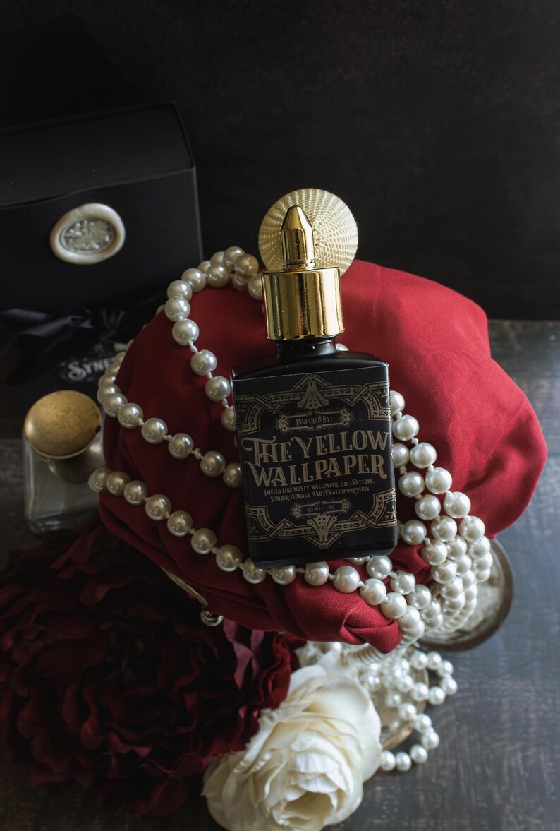 A bottle of “The Yellow Wallpaper” perfume with a gold atomizer sits atop deep red fabric, draped in pearls and surrounded by dark red and white roses. The composition, dramatic with shadows and heavy textures, evokes a sense of trapped opulence—luxury slipping into confinement, as if the beauty itself carries a fevered unease.