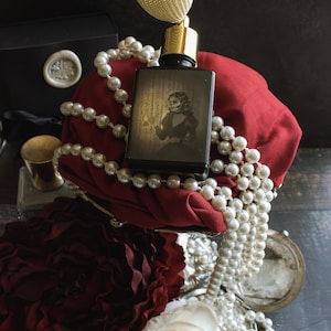 A bottle of “The Yellow Wallpaper” perfume with a golden atomizer rests against red fabric, draped in pearl strands, its illustrated label depicting a spectral woman holding a lantern. The dimly lit scene, framed by wilted roses and shadow, feels like a fever dream of beauty unraveling—where opulence curdles into claustrophobic dread.
