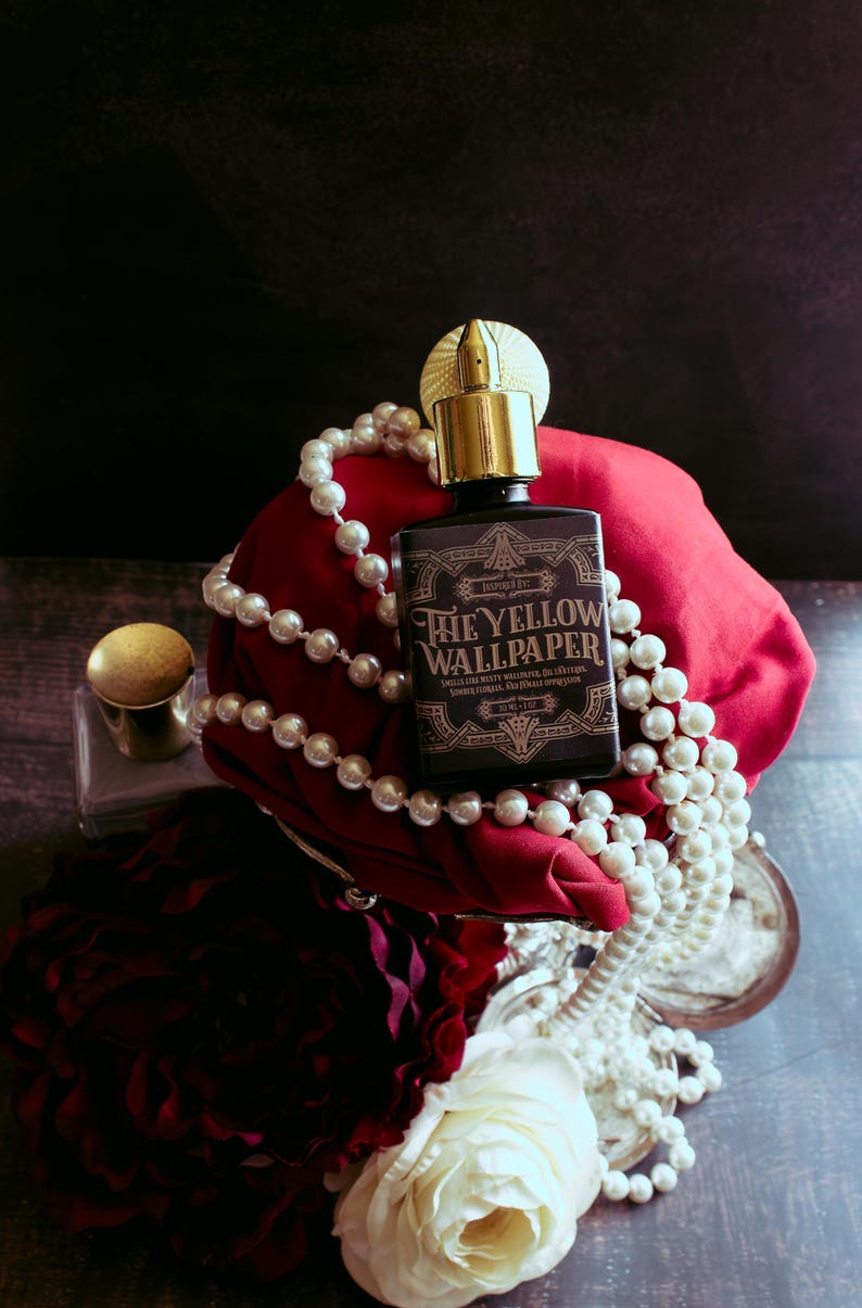 A 30ml perfume bottle labeled “The Yellow Wallpaper” with a gold atomizer rests on rich red fabric, draped in strands of pearls and framed by white and dark red roses. The scene is staged like a decadent still life, yet the dim lighting and heavy shadows lend a feverish, claustrophobic mood—an atmosphere of beauty fraying at the edges of madness.