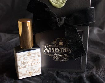 Op de afbeelding: Een klein flesje parfum met een gouden dop en een wit etiket met de tekst "Midsummer Night's Dream". Het flesje staat op een zwarte stoffen ondergrond naast een zwarte doos met een gouden waszegel en een zwart fluwelen lint. De doos heeft een wit etiket met de tekst "Synesthes Scent Co."