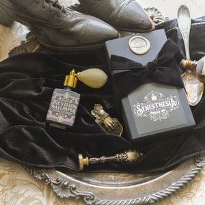 A bottle of The Yellow Wallpaper perfume with a gold atomizer rests in a silver tray alongside its Synesthesia Scent Co. gift box, draped in black velvet and surrounded by antique trinkets and scuffed Victorian boots. The composition evokes a relic unearthed from a haunted parlor, steeped in faded grandeur and the unsettling whispers of history.