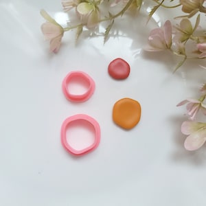 May include: A selection of polymer clay pieces and pink cookie cutters. Two pink, irregular-shaped cutters are next to two clay pieces, one red and one orange. The clay pieces are small, measuring approximately 2 cm in diameter.