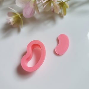 Puede incluir: Dos cortadores de galletas rosas en forma de riñón. Uno es una forma sólida y el otro es un contorno hueco. Los cortadores están sobre una superficie blanca, con flores rosas en el fondo.