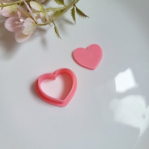 May include: Two pink heart-shaped cookie cutters, one with a cut-out centre and one solid, on a white surface.