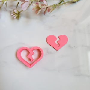 Peut inclure: Deux objets roses en forme de cœur, un entier et un brisé, sont présentés sur une surface blanche. Le cœur brisé présente une fissure en zigzag. De délicates fleurs roses sont en arrière-plan.