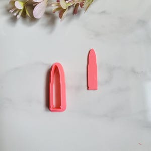 Pode incluir: Duas cortadores de biscoitos cor-de-rosa em forma de bala. Um é uma bala sólida, o outro um contorno oco. Os cortadores são de plástico e estão sobre uma superfície de mármore branco.