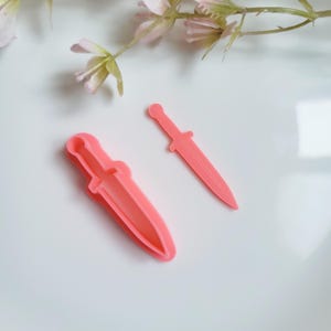 May include: Two pink plastic cookie cutters shaped like a dagger. The cutters are on a white surface.
