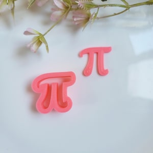 Könnte beinhalten: Zwei rosafarbene Plastik-Ausstecher in Form des mathematischen Symbols Pi. Die Ausstecher befinden sich auf einer weißen Oberfläche.