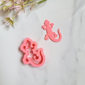 Könnte beinhalten: Zwei rosa, eidechsenförmige Gegenstände. Einer ist eine feste Eidechse, und der andere ist ein Ausstecher in Eidechsenform. Die Gegenstände befinden sich auf einer weißen Marmoroberfläche mit einem Zweig rosa Blüten in der oberen linken Ecke.