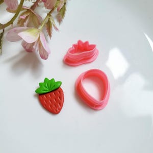 May include: A set of three pink plastic cookie cutters shaped like a strawberry. The cutters are on a white surface.