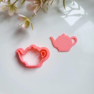 May include: Two pink plastic teapot cookie cutters. The larger cutter is solid and the smaller cutter is an outline.