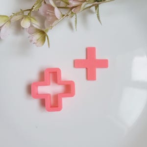 May include: Two pink cross-shaped cookie cutters, one with a solid outline and one solid, sit on a white surface. Delicate pink flowers and greenery are in the upper left corner. The cutters are likely for baking or crafting.
