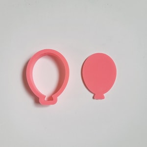 May include: Two pink plastic cookie cutters shaped like balloons. One cutter is a solid shape, the other is an outline.